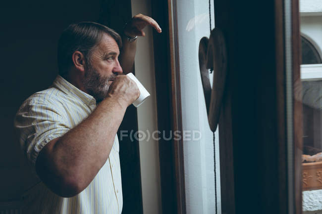 Продуманий старший чоловік має каву, дивлячись крізь вікно вдома — стокове фото
