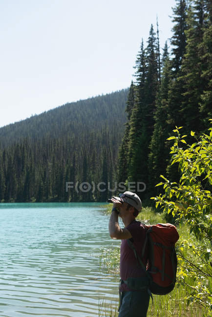 Male hiker looking through binoculars near riverside in mountains — Stock Photo