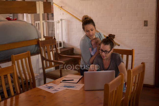 Pareja lesbiana discutiendo sobre el ordenador portátil mientras sostiene al bebé en casa - foto de stock