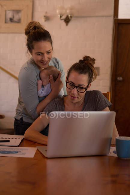 Pareja lesbiana usando portátil mientras sostiene al bebé en casa - foto de stock