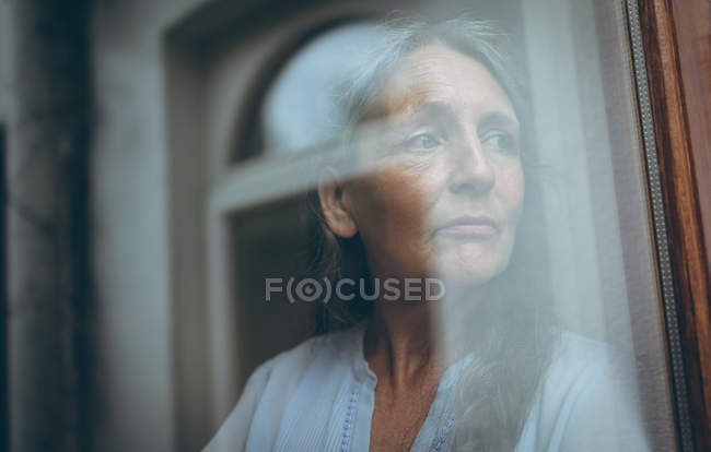 Nachdenkliche Seniorin schaut zu Hause durch Fenster — Stockfoto