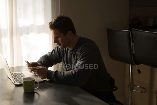 Hombre usando el teléfono móvil en la mesa de comedor en casa - foto de stock