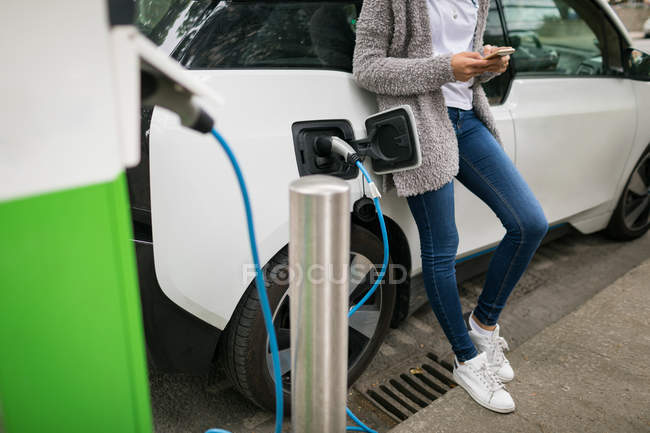 Section basse de la femme utilisant un téléphone portable tout en rechargeant la voiture électrique à la station de charge — Photo de stock