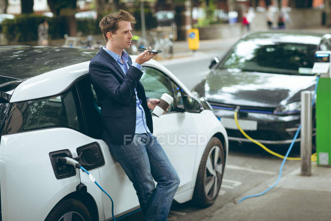 Empresário falando no telefone celular enquanto carrega carro elétrico na estação de carregamento — Fotografia de Stock