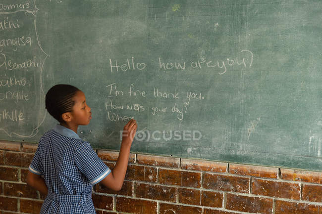 Schoolgirl writing on chalkboard in classroom at school — Stock Photo