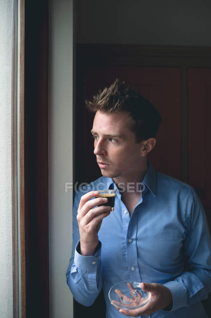 Задумчивый человек смотрит в окно, когда пьет кофе дома — стоковое фото
