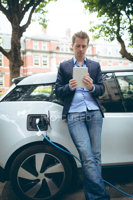 Homme d'affaires utilisant une tablette numérique tout en rechargeant la voiture électrique à la station de charge — Photo de stock
