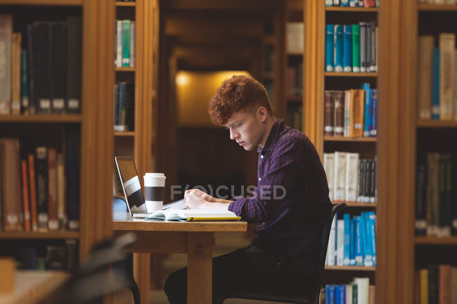 Vista lateral del estudiante universitario leyendo un libro en la biblioteca - foto de stock