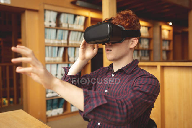 Joven estudiante universitario usando auriculares de realidad virtual en la biblioteca - foto de stock
