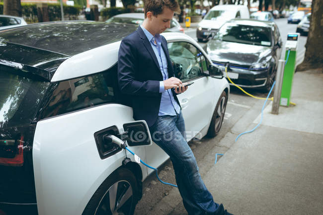 Homme d'affaires utilisant un téléphone portable tout en rechargeant la voiture électrique à la station de charge — Photo de stock