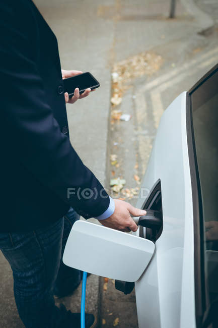 Vue grand angle de l'homme d'affaires utilisant un téléphone portable tout en rechargeant la voiture électrique à la station de charge — Photo de stock