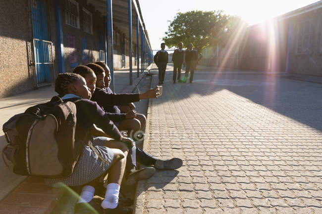 Studenti che si scattano selfie con il cellulare nel campus scolastico in una giornata di sole — Foto stock