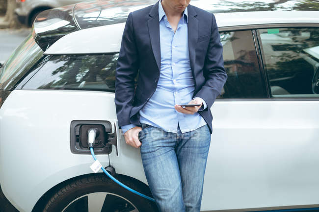 Seção média do homem de negócios usando telefone celular ao carregar carro elétrico na estação de carregamento — Fotografia de Stock