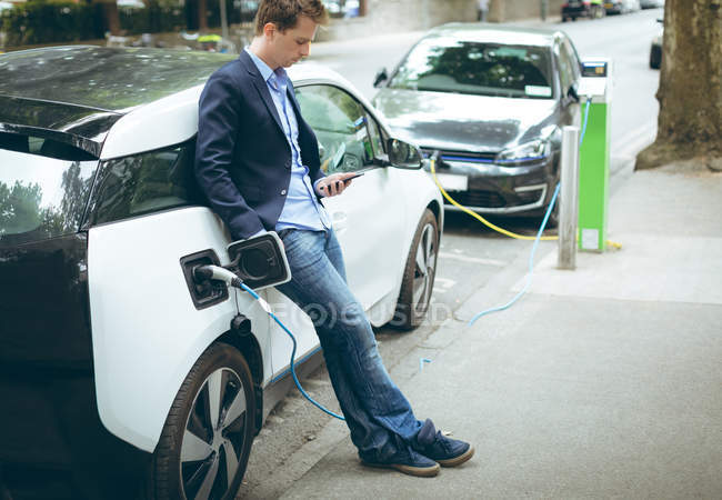 Homme d'affaires utilisant un téléphone portable tout en rechargeant la voiture électrique à la station de charge — Photo de stock
