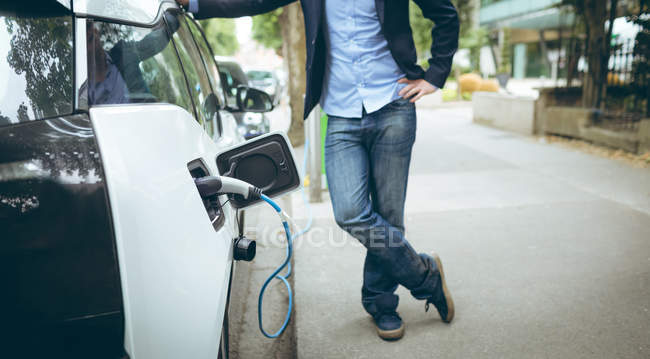 Sección baja del hombre de negocios que carga el coche eléctrico en la estación de carga - foto de stock