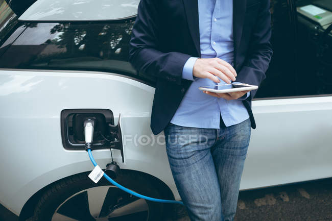 Homme d'affaires utilisant une tablette numérique tout en rechargeant la voiture électrique à la station de charge — Photo de stock