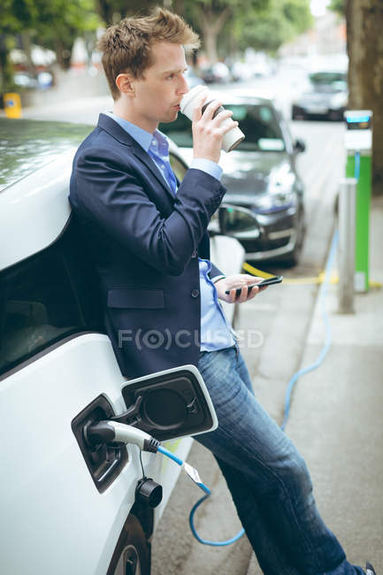 Бизнесмен пьет кофе во время зарядки электромобиля на зарядной станции — стоковое фото
