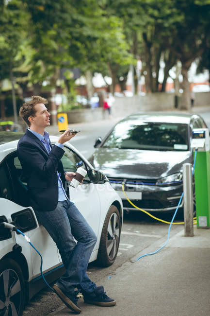 Homme d'affaires parlant sur un téléphone portable tout en rechargeant une voiture électrique à la station de charge — Photo de stock