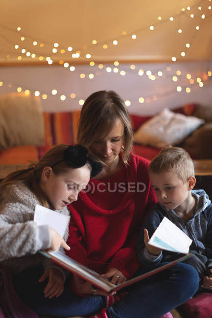 Mère et enfants regardant un album photo à la maison contre les lumières de Noël — Photo de stock