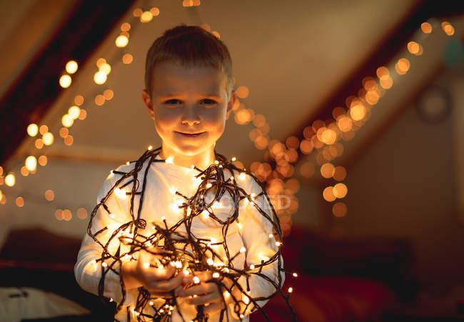 Sorrindo menino segurando luzes de fadas iluminadas em casa — Fotografia de Stock