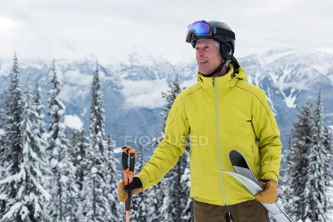 Hombre mayor sonriente con bastón de esquí y tabla de esquí de pie en la región nevada - foto de stock