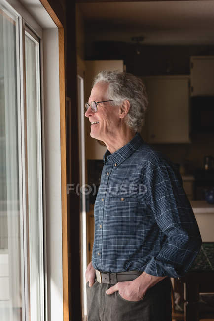 Hombre mayor sonriente mirando a través de la ventana en casa - foto de stock