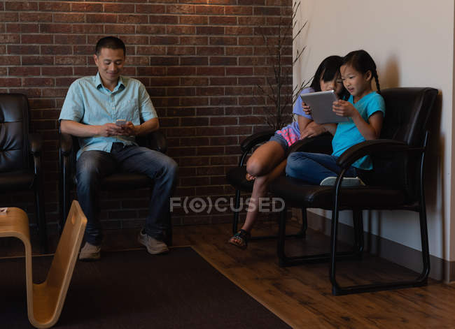 Padre utilizzando il telefono cellulare mentre le figlie utilizzando tablet digitale in clinica dentale — Foto stock