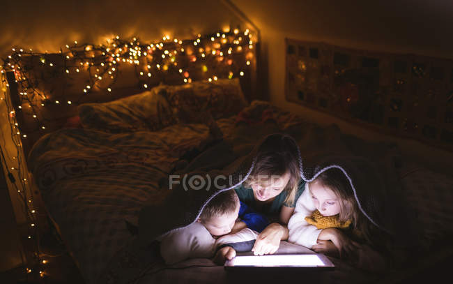 Madre e hijos debajo de la manta usando tableta digital contra la decoración de Navidad - foto de stock