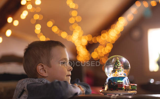 Niño curioso mirando el árbol de Navidad bola de nieve en casa - foto de stock