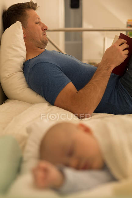 Bebé niño durmiendo mientras padre leyendo libro en el dormitorio en casa - foto de stock