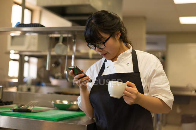 Жіночий шеф-кухар використовує мобільний телефон, маючи каву на кухні — стокове фото