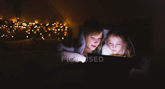Mère et fille sous la couverture en utilisant une tablette numérique contre les lumières de Noël — Photo de stock