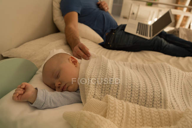 Bebé niño durmiendo mientras el padre usando el ordenador portátil en el dormitorio en casa - foto de stock
