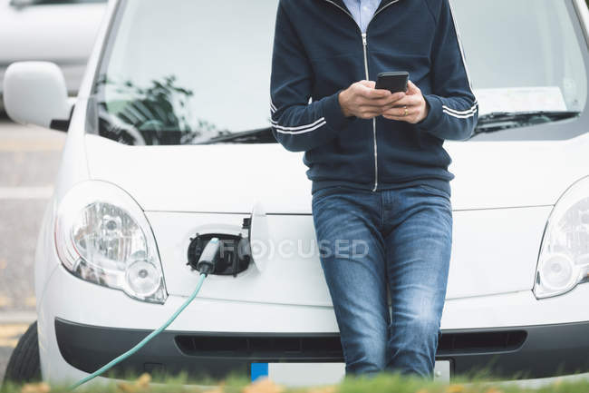 Partie médiane de l'homme utilisant un téléphone portable tout en rechargeant la voiture électrique à la station de charge — Photo de stock