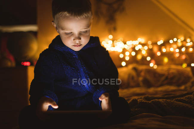 Menino concentrado em casaco azul usando tablet digital contra luzes de Natal — Fotografia de Stock