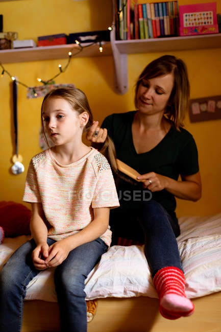 Madre peinando hijas cabello en la cama en casa - foto de stock
