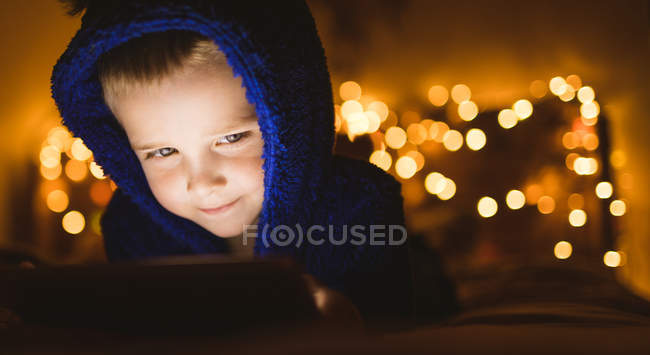 Primer plano del niño con chaqueta azul usando tableta digital contra luces navideñas - foto de stock