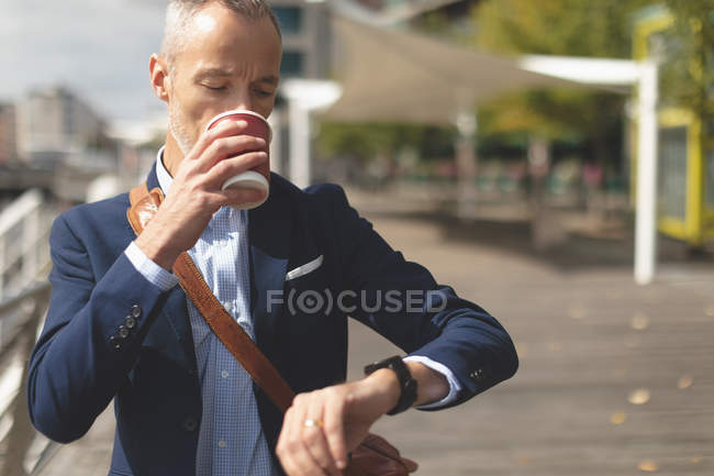 Geschäftsmann trinkt Kaffee, während er die Uhrzeit auf der Smartwatch an der Promenade kontrolliert — Stockfoto