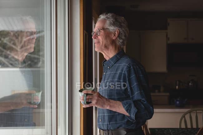 Homem sênior olhando pela janela enquanto toma café em casa — Fotografia de Stock