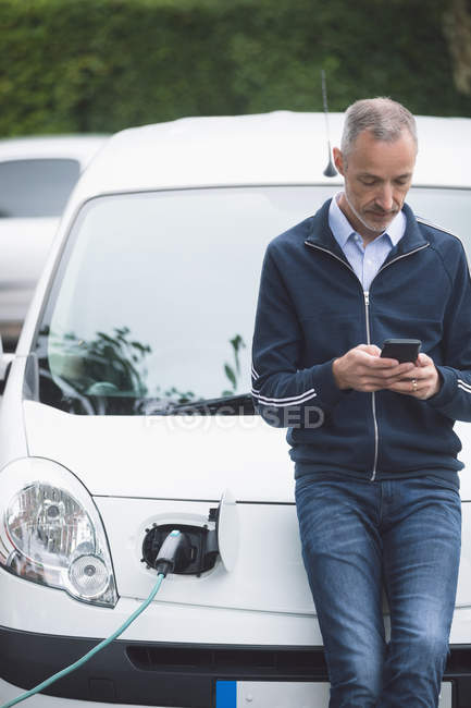 Homem usando telefone celular ao carregar carro elétrico na estação de carregamento — Fotografia de Stock