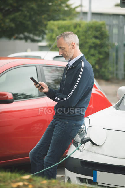 Hombre usando el teléfono móvil mientras carga el coche eléctrico en la estación de carga - foto de stock
