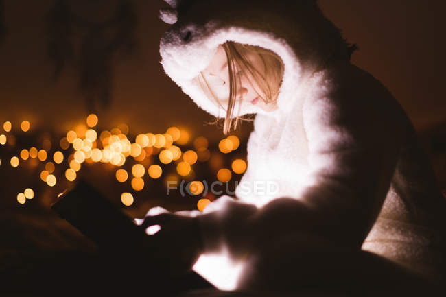 Chica en chaqueta con capucha utilizando tableta digital contra las luces de Navidad - foto de stock