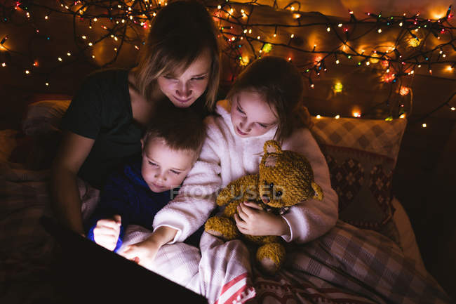 Mutter und Kinder mit digitalem Tablet im Bett gegen Weihnachtsdekoration — Stockfoto