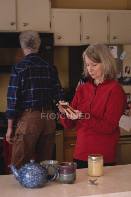 Donna anziana che utilizza il telefono cellulare mentre l'uomo lavora in background in cucina — Foto stock