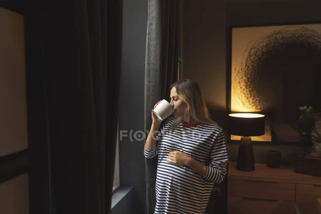 Беременная женщина пьет кофе у окна дома — стоковое фото