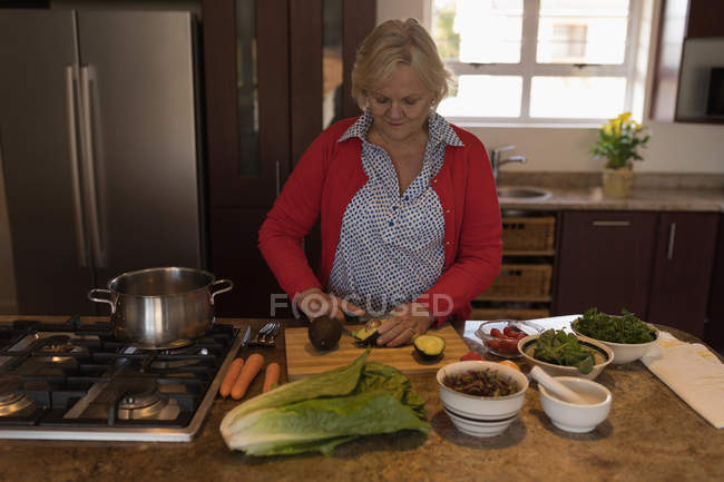 Старшая женщина режет авокадо на кухне дома — стоковое фото