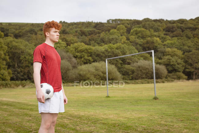 Joven jugador de fútbol de pie con pelota de fútbol en el campo - foto de stock