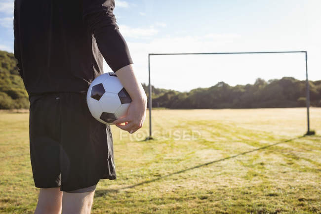 Parte média do jogador de futebol de pé com bola de futebol no campo — Fotografia de Stock