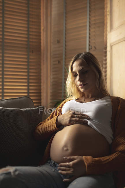 Thoughtful pregnant woman sitting on sofa touching her belly — Stock Photo