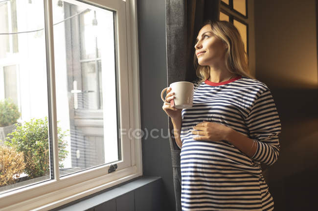 Schwangere mit Kaffeetasse steht zu Hause am Fenster — Stockfoto